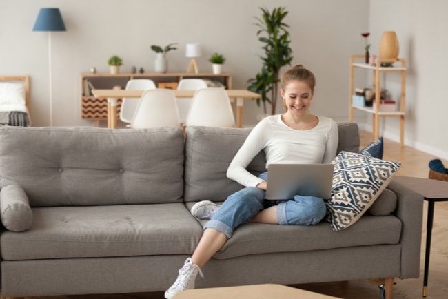 leende ung kvinna med laptop i soffa i sin lägenhet