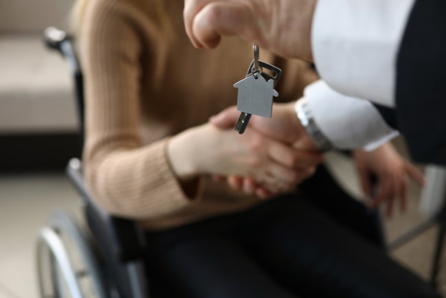 Person räcker skakar en rullstolsbunden kvinnas hand och räcker över nyckeln till ett boende.