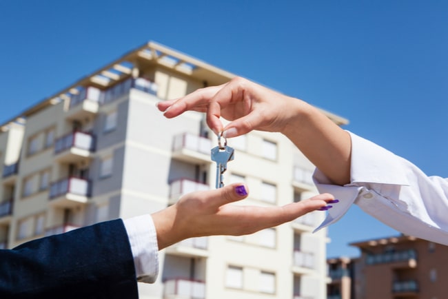 Närbild på hand som överlämnar nycklar till en annan hand med bostadshus i bakgrunden.